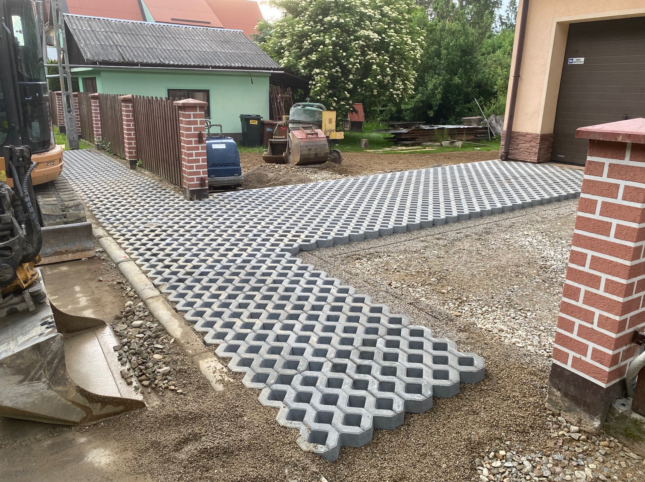 Ułożona ażurowa kostka betonowa na podjeździe w Niedzicy, widoczny fragment koparki, ogrodzenie z ceglanymi słupkami i budynek gospodarczy w tle.