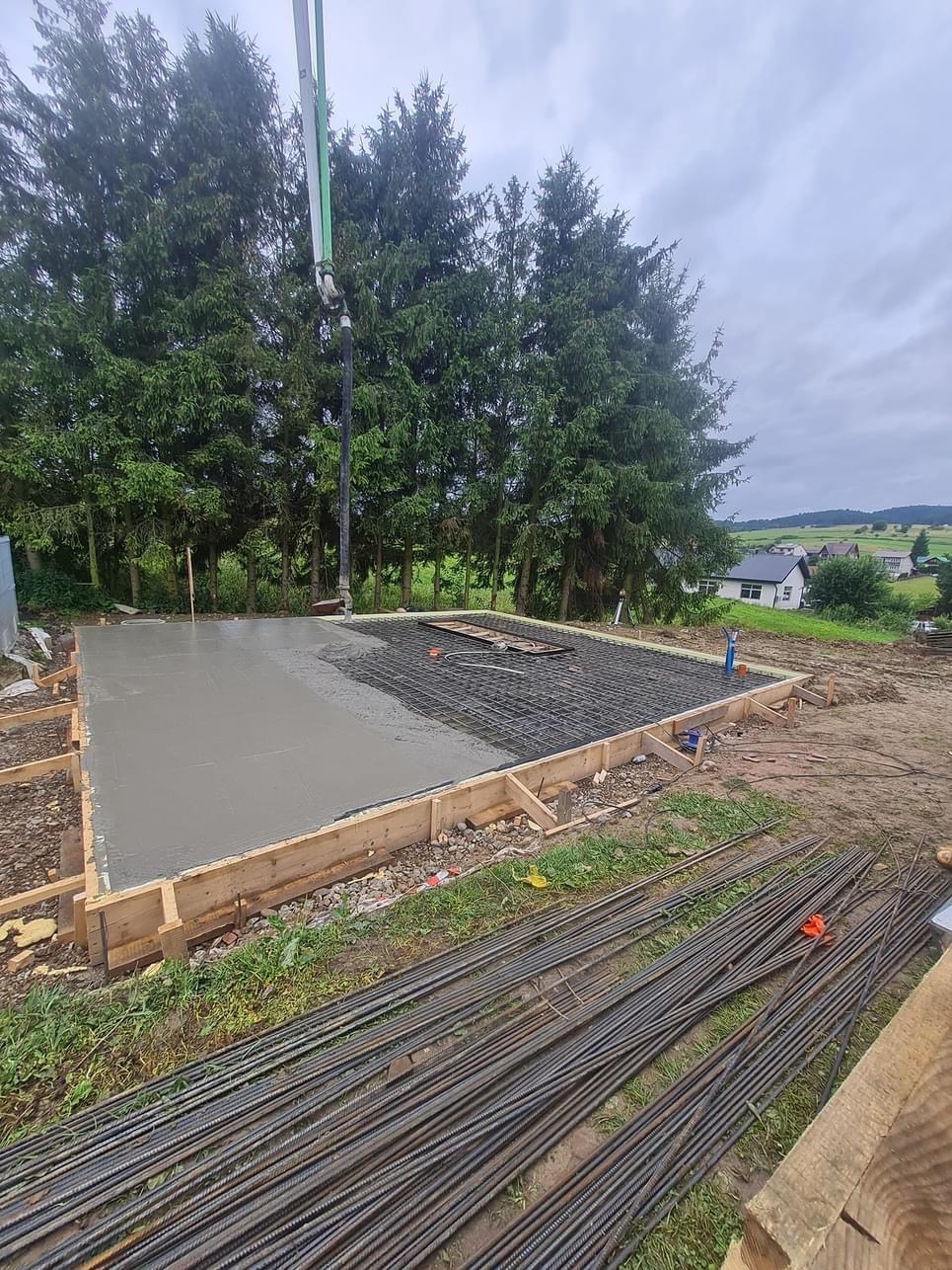 Wylewanie fundamentów pod budynek z użyciem pompy do betonu. Część fundamentu zalana, widoczne zbrojenie, szalunek drewniany i stos prętów zbrojeniowych na ziemi. W tle drzewa i zabudowania.