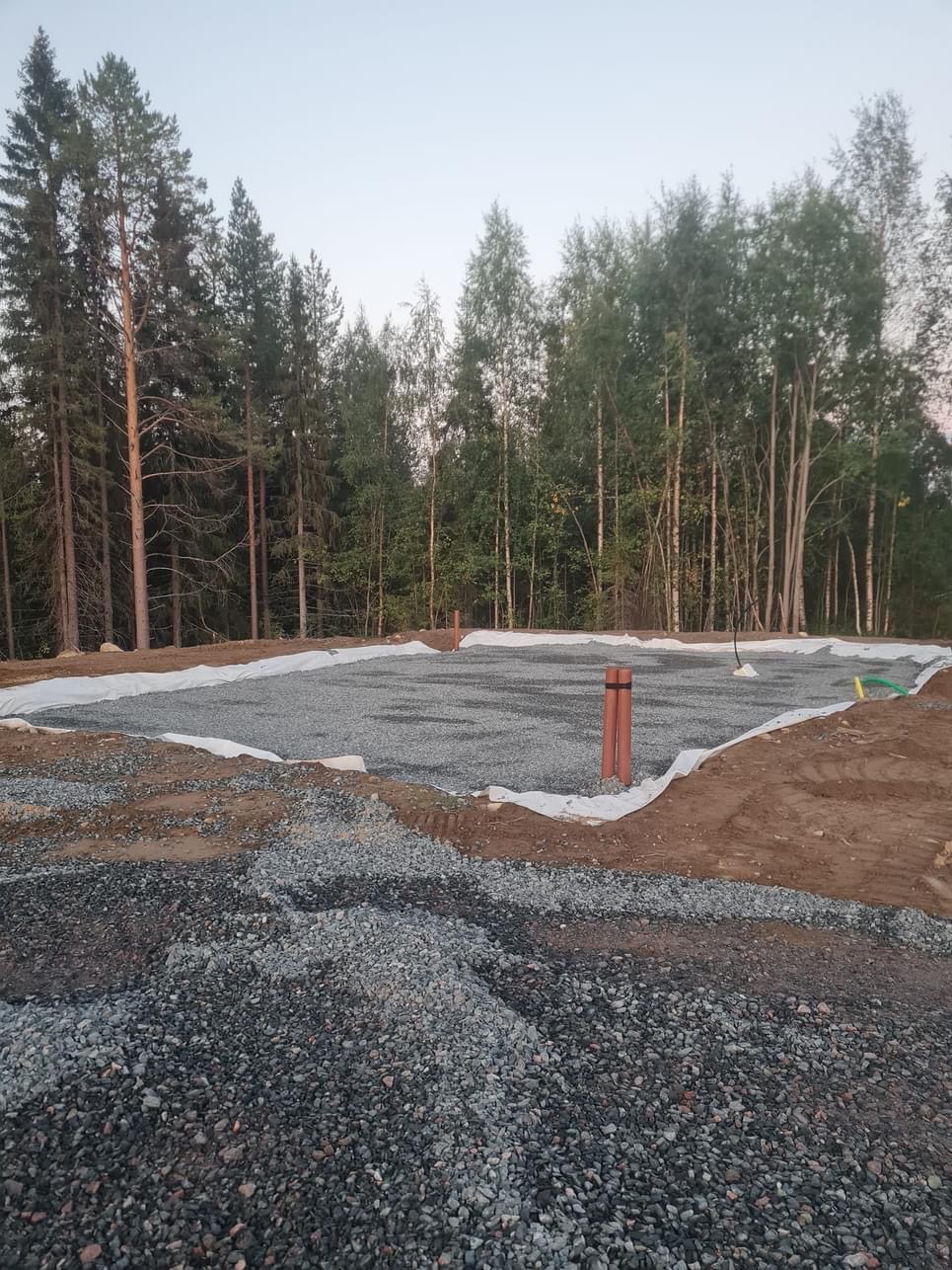 Przygotowany teren pod budowę: warstwa geowłókniny przykryta żwirem, widoczne rury kanalizacyjne, otoczenie leśne, w tle drzewa.