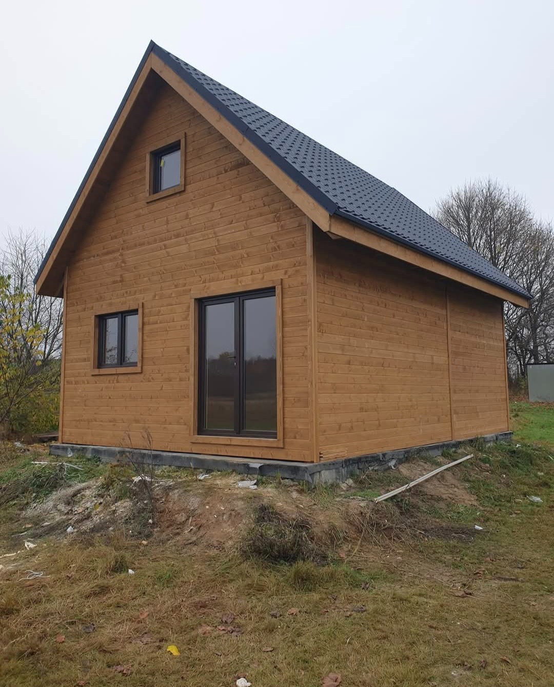 Surowy, drewniany budynek z ciemną, spadzistą dachówką, usytuowany na nierównym terenie z widoczną trawą i drzewami w tle.