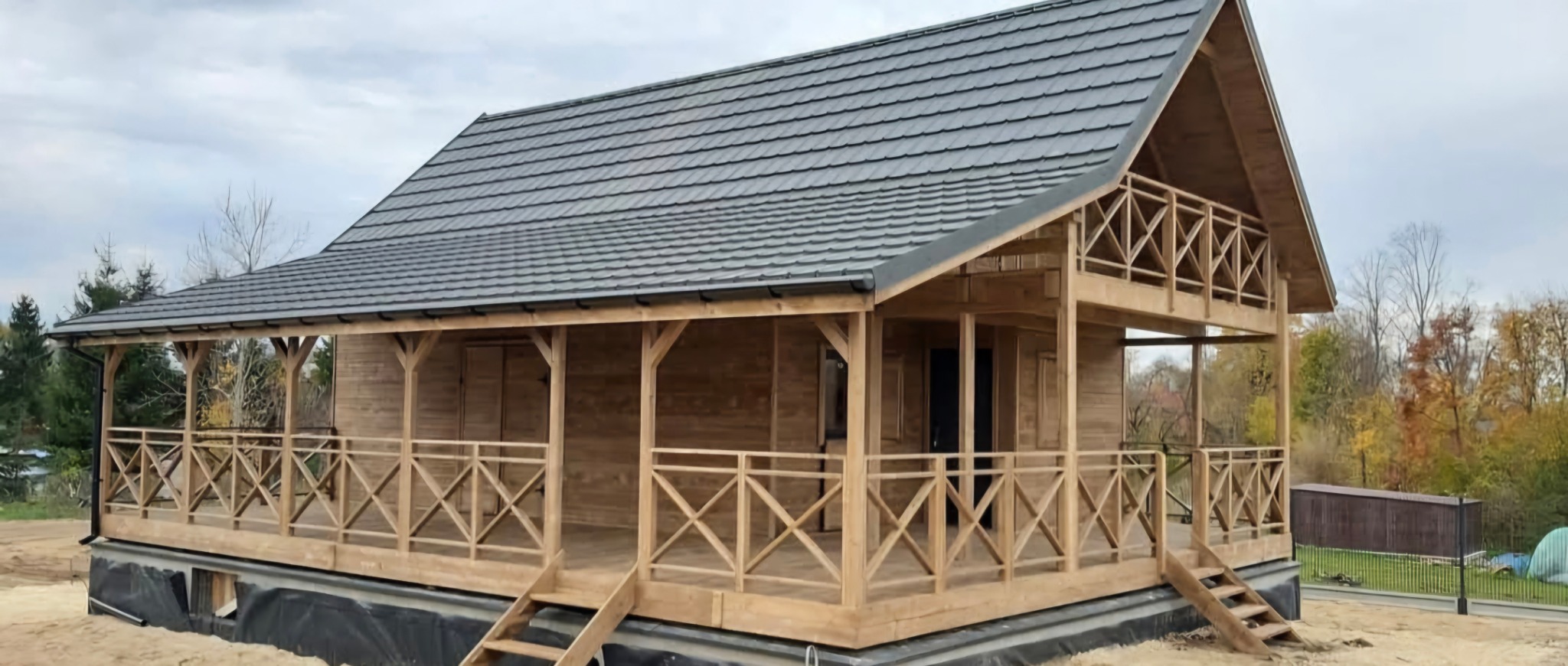 Niski, parterowy budynek z drewna z ciemnoszarą dachówką, otoczony werandą z drewnianą balustradą w kształcie krzyży, posadowiony na fundamencie z czarną izolacją, na tle jesiennego krajobrazu.