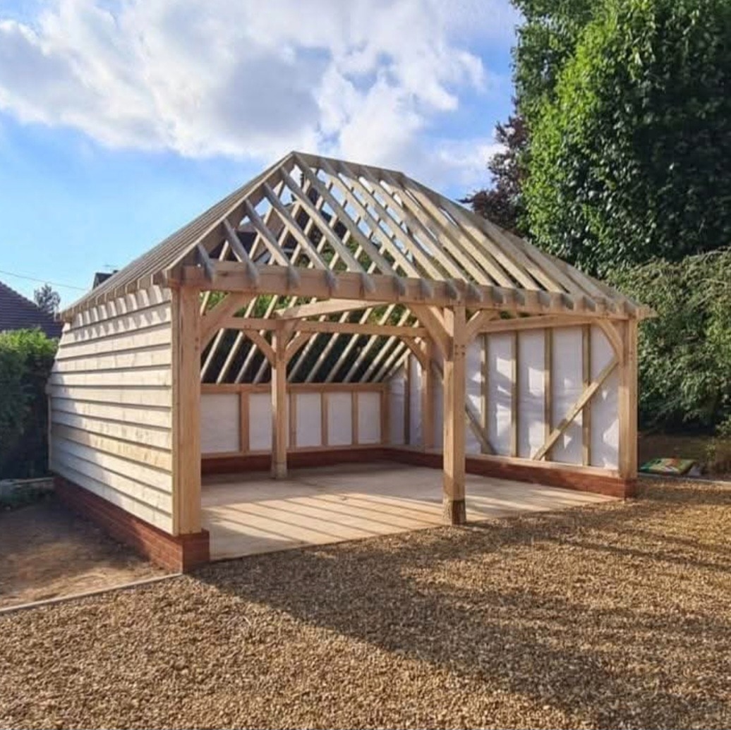 Konstrukcja drewniana z elementami szkieletowymi, częściowo obłożona deskami, na fundamencie z cegieł, otoczona żwirem i zielenią.