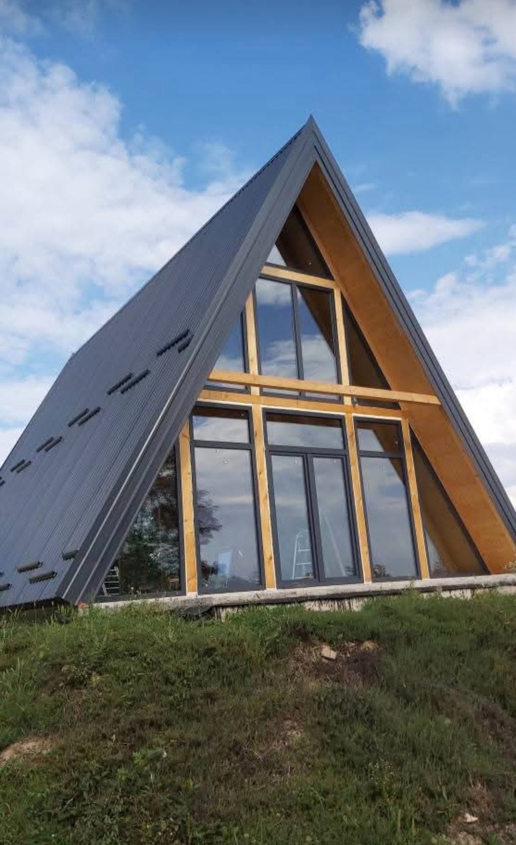 Nowoczesny dom szkieletowy w kształcie litery A z ciemnoszarą blachą na dachu, dużymi oknami i drewnianą konstrukcją, na tle błękitnego nieba z chmurami.