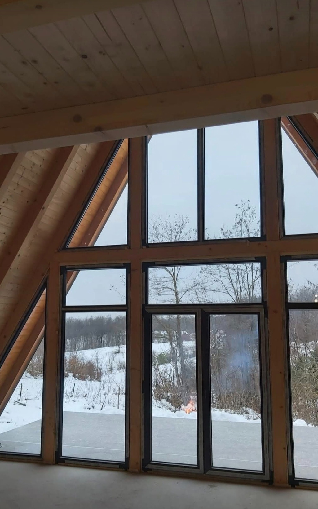 Wnętrze surowego domu w konstrukcji szkieletowej z widokiem na zimowy krajobraz przez duże okna; na zewnątrz, ognisko.