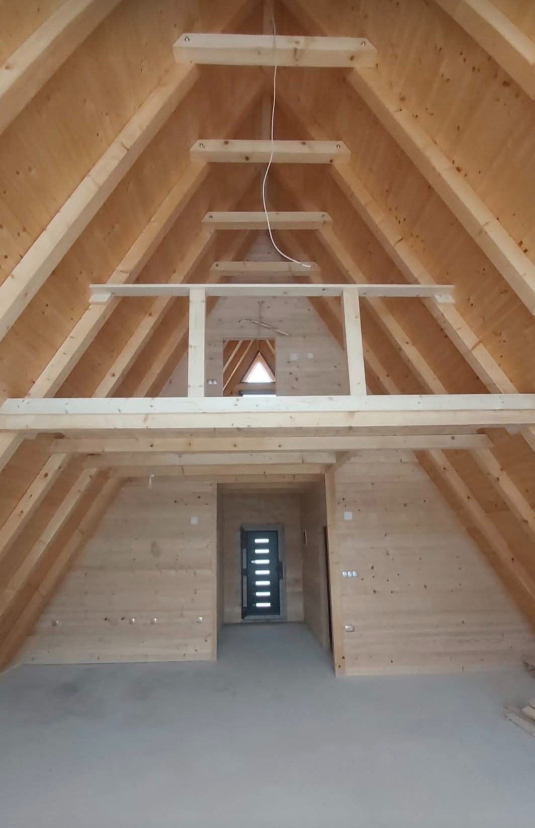 Wnętrze w stanie surowym otwartym: widok na drewnianą konstrukcję dachu, antresolę z barierką i drzwi wejściowe w budynku z drewnianych paneli.