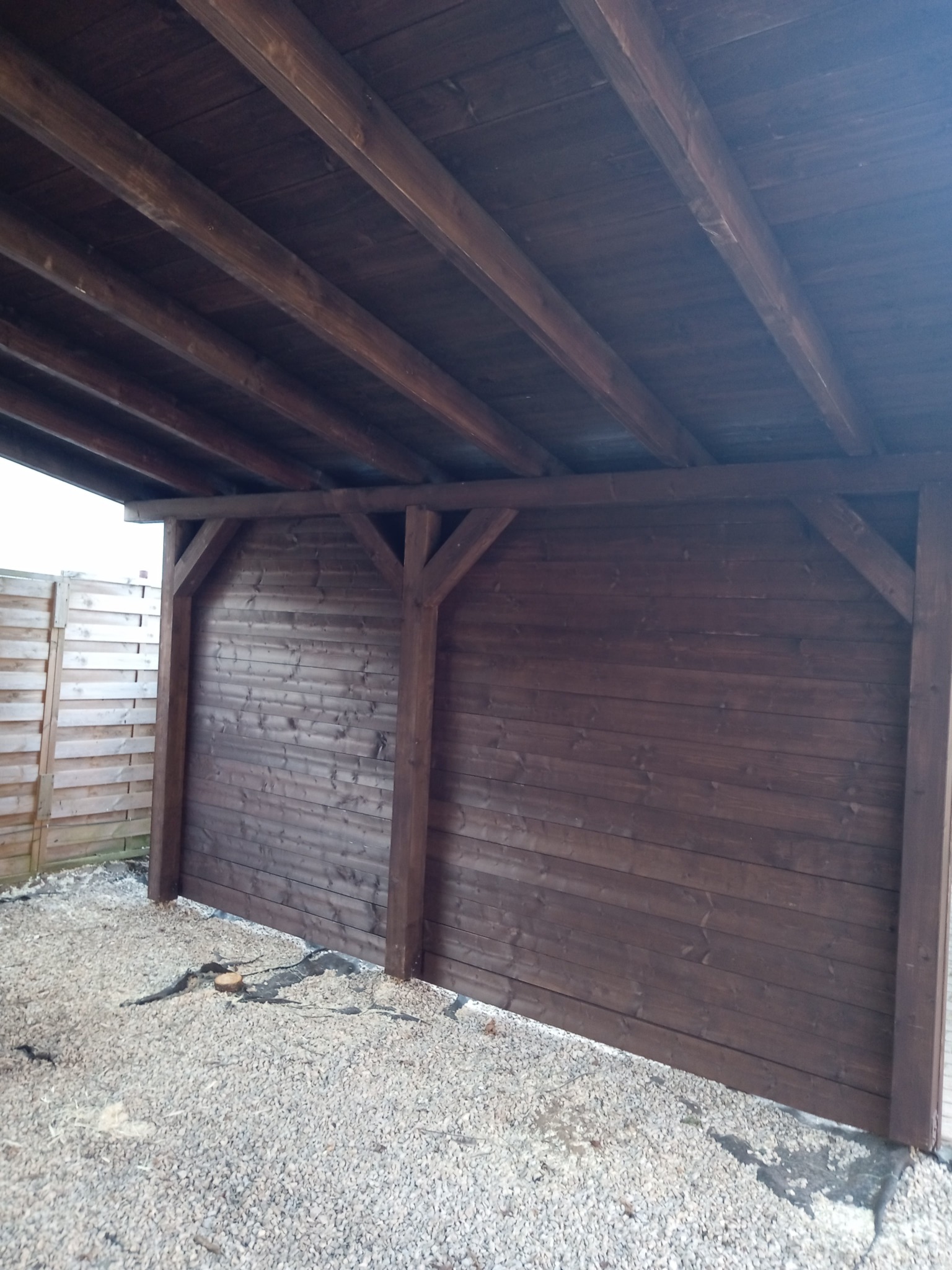 Drewniana wiata z ciemnobrązowych desek, zadaszona, z podłożem wysypanym drobnym kamieniem.