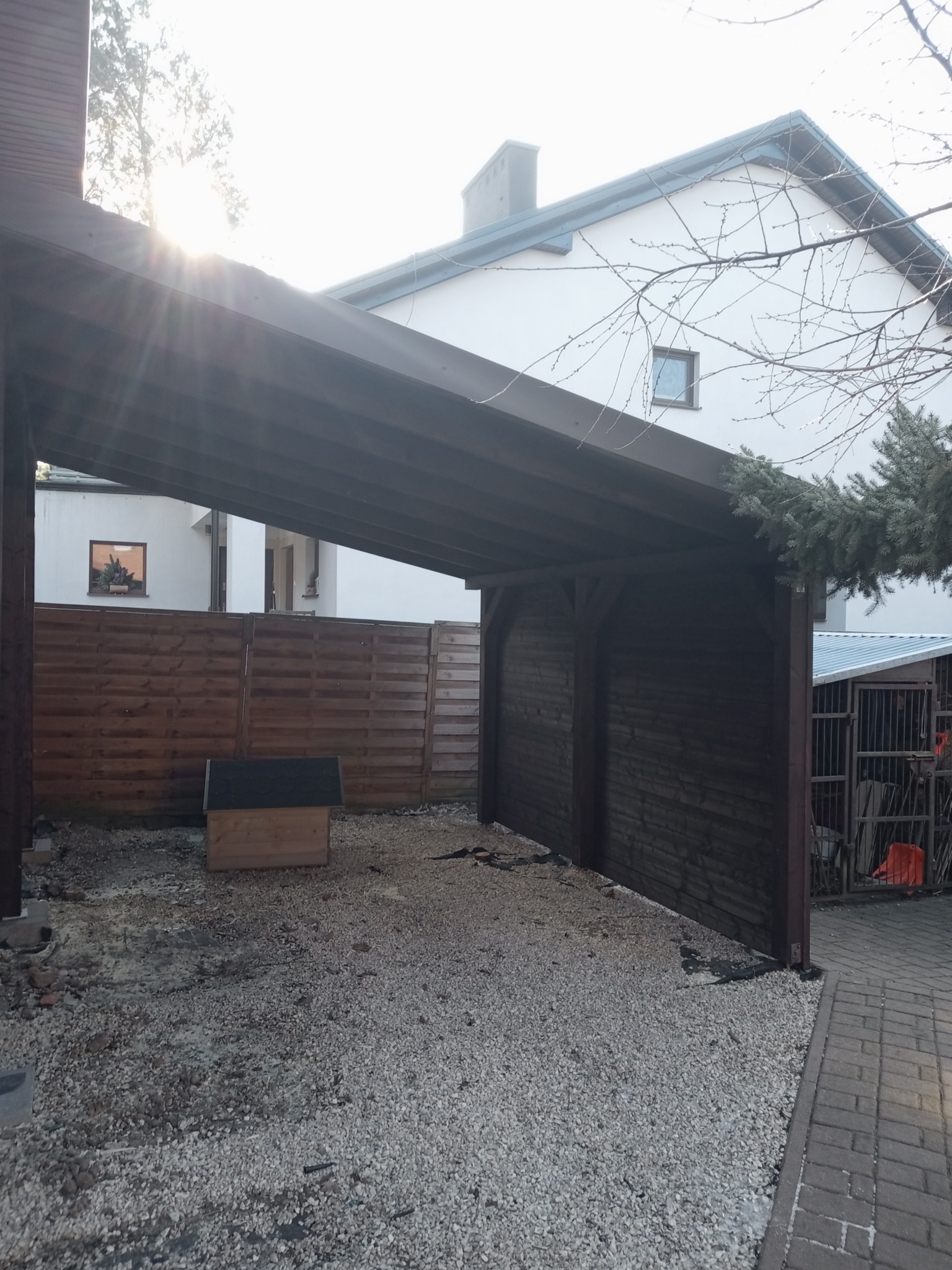 Drewniana wiata garażowa z ciemnego drewna, widoczna z zewnątrz, z małą budą dla psa w pobliżu, na tle domu z białymi ścianami i drewnianym płotem.