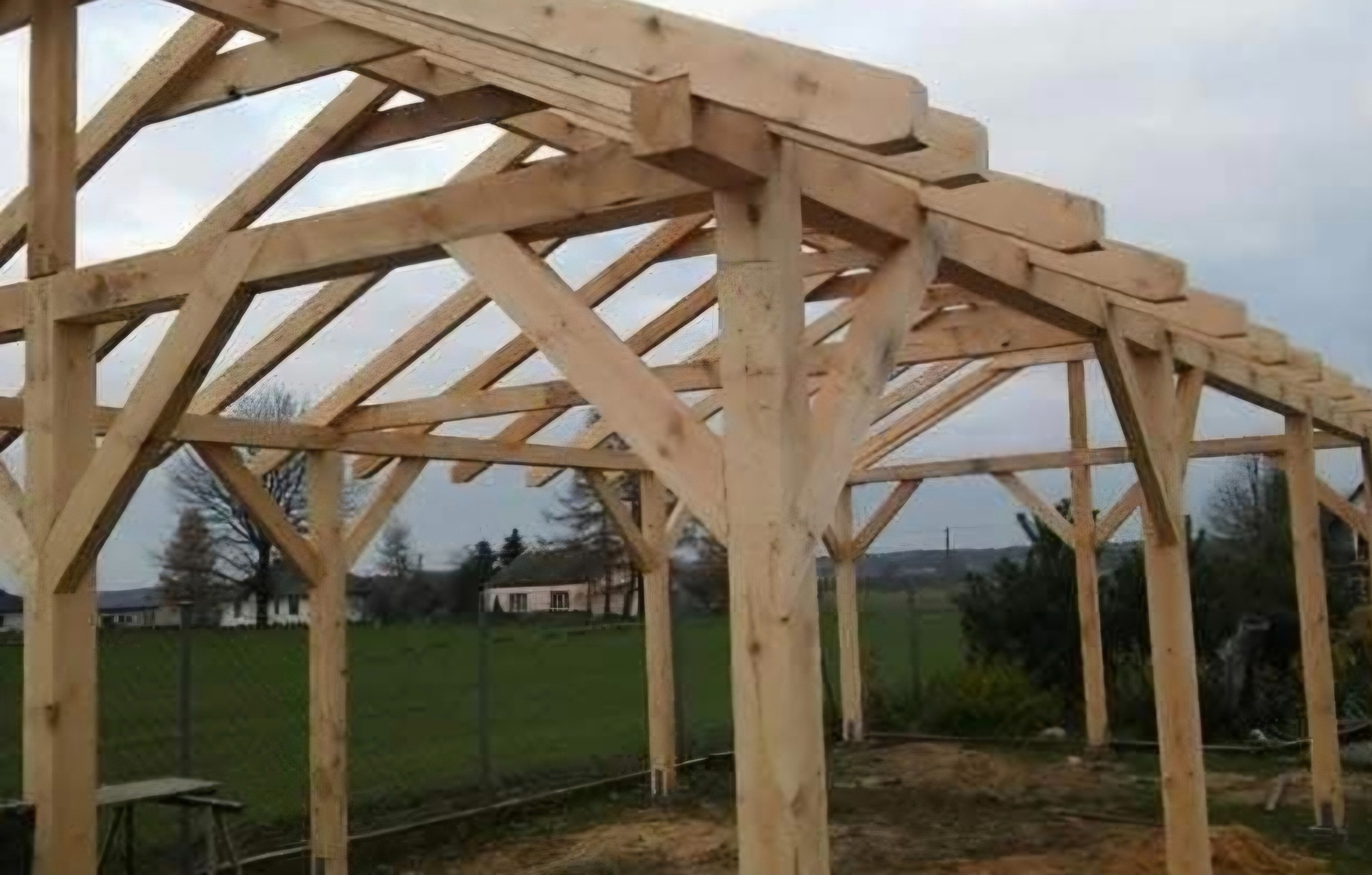 Surowa konstrukcja domu szkieletowego w Kosowie, widoczne drewniane elementy nośne i więźba dachowa na tle zieleni.