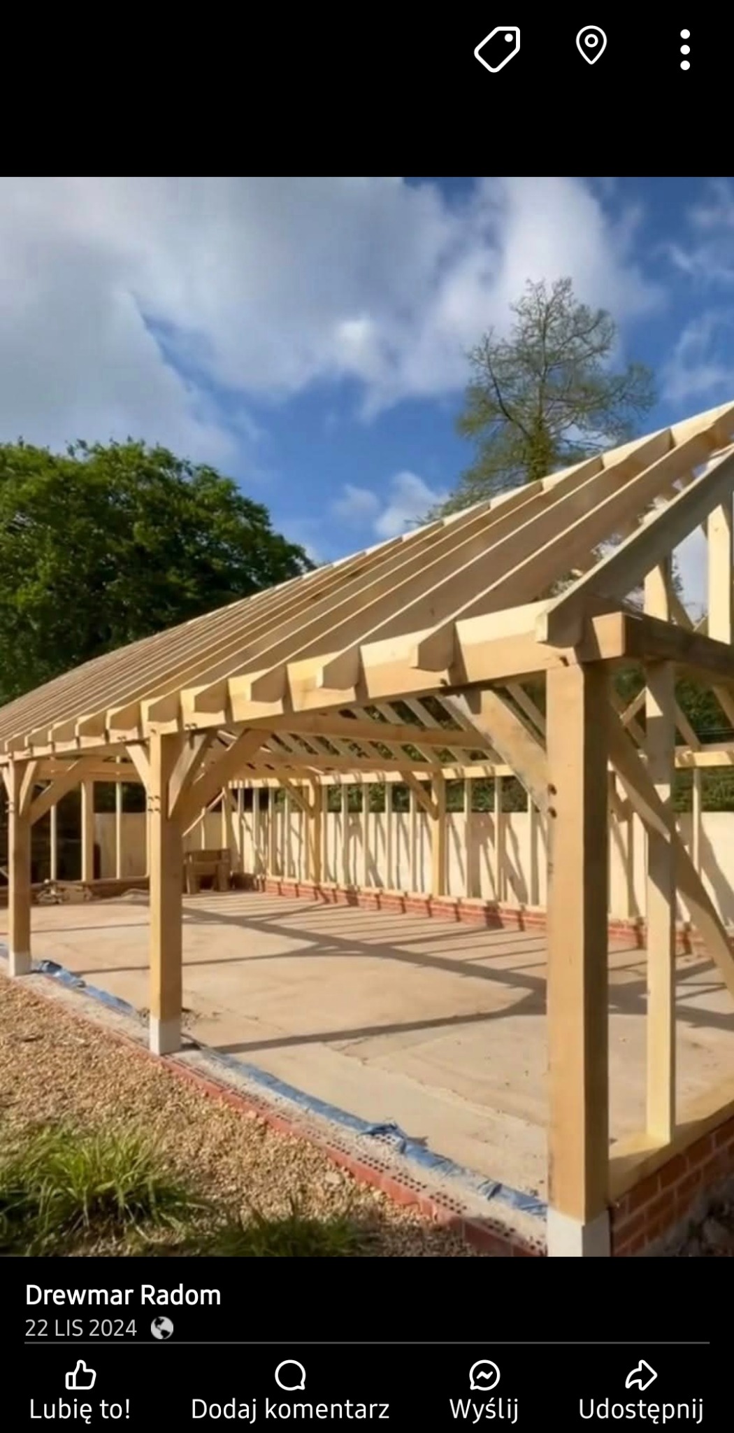 Konstrukcja drewniana z belek i krokwi w trakcie budowy, widoczne fundamenty z cegieł, tło z zielenią i błękitnym niebem.