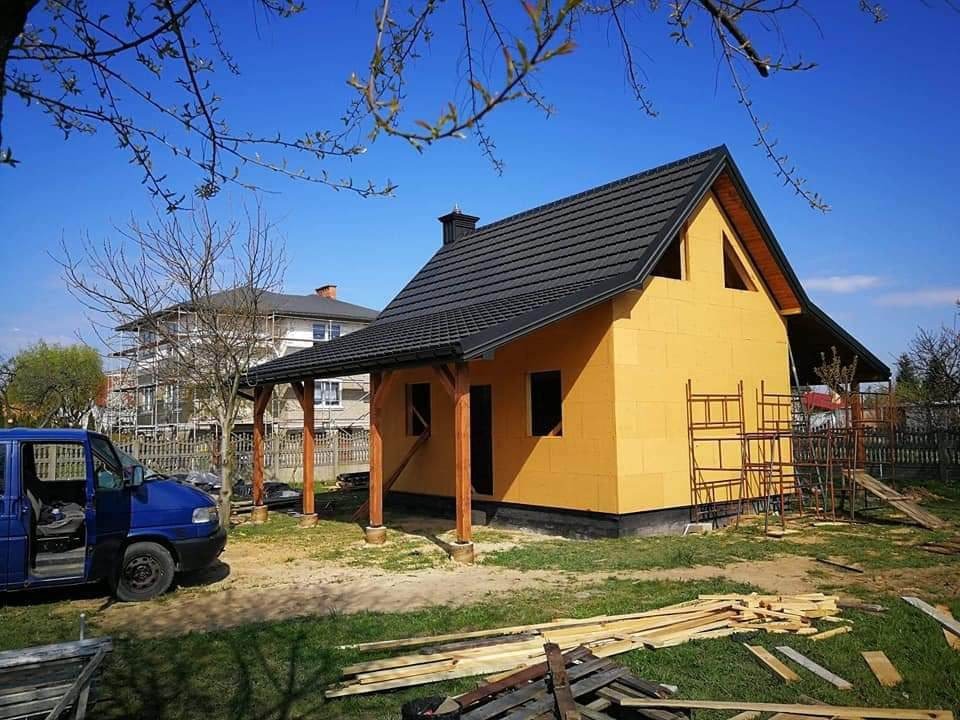 Budowa domu w technologii szkieletowej: surowe ściany w kolorze żółtym, czarny dach, drewniana konstrukcja werandy i rusztowania w tle, widoczny niebieski samochód dostawczy.