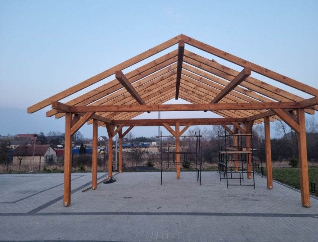 Konstrukcja drewniana zadaszenia w trakcie budowy, widoczne rusztowania i otoczenie z budynkami w tle, sfotografowane o zmierzchu.