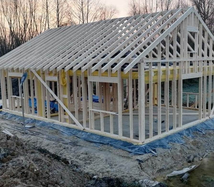 Konstrukcja szkieletowa domu jednorodzinnego w trakcie budowy, widoczne drewniane elementy nośne, żółta folia izolacyjna i fundament zabezpieczony niebieską folią, w tle drzewa bez liści.