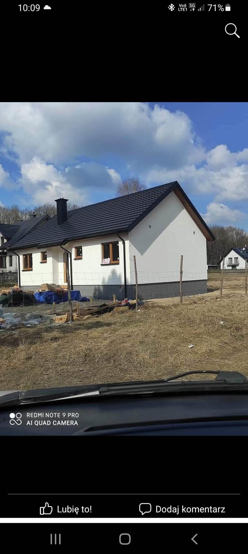 Niedokończony budynek z białymi ścianami i ciemnym dachem, widoczny zza szyby samochodu, teren wokół w trakcie przygotowywania.