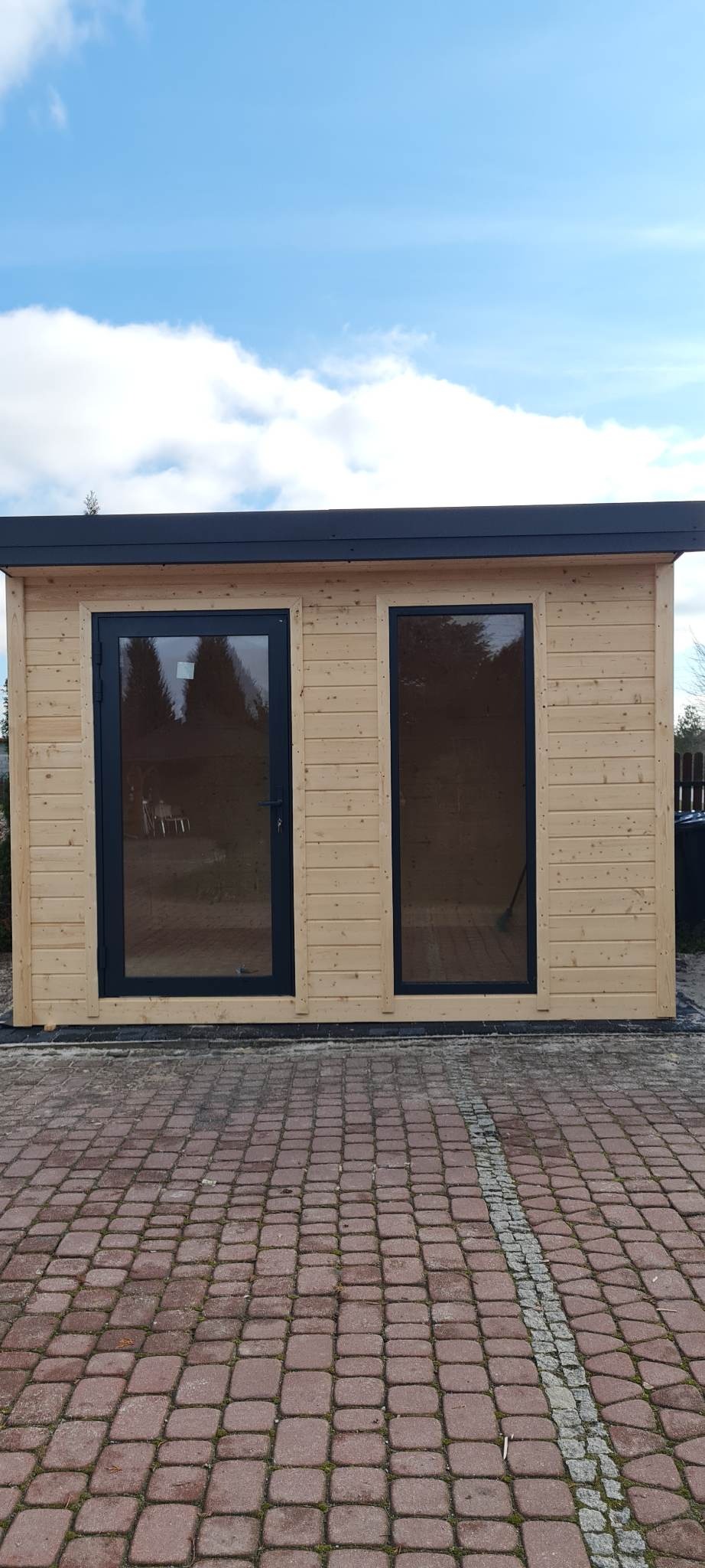 Niewielki drewniany budynek z dwoma ciemnymi oknami i drzwiami, postawiony na czerwonej kostce brukowej, widziany w słoneczny dzień.