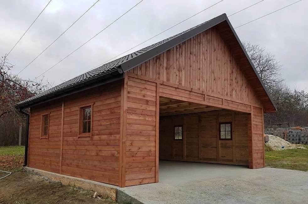 Drewniany garaż z elementami konstrukcji szkieletowej, wykończony na zewnątrz brązową boazerią, z widocznym betonowym podjazdem i stertą kostki brukowej w tle.