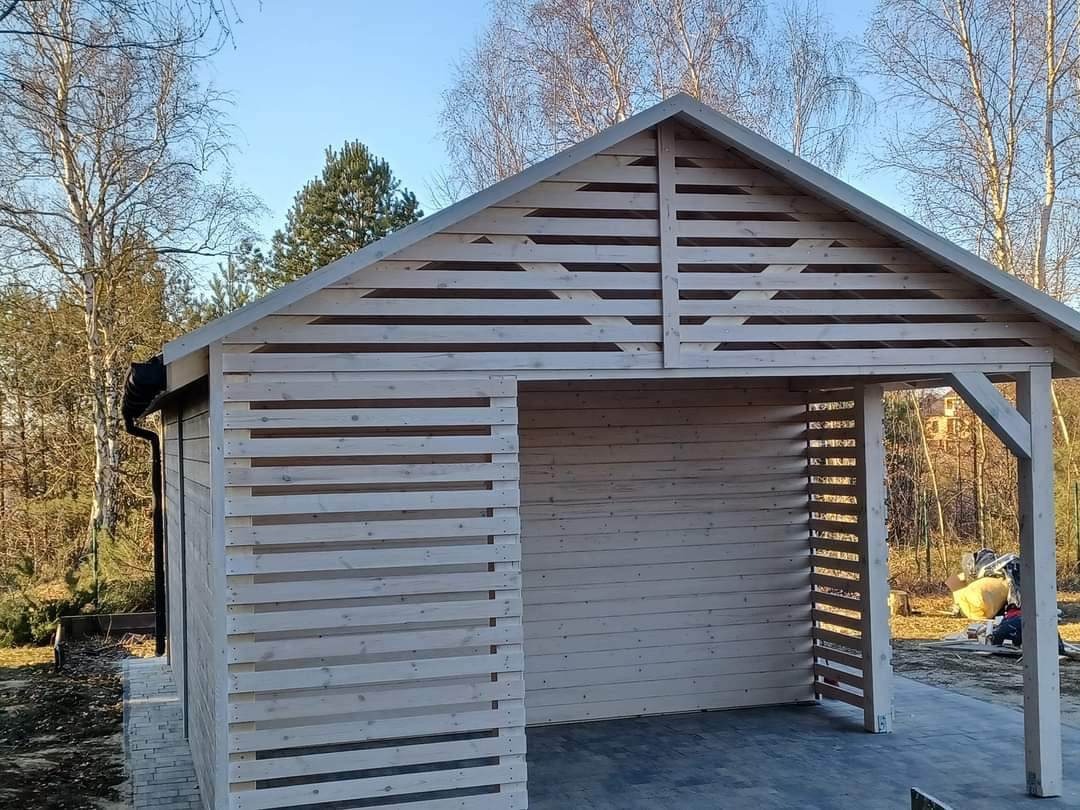 Surowy, drewniany dom szkieletowy z poziomymi listwami, zadaszona wiata garażowa z widoczną konstrukcją nośną, na tle drzew bezlistnych.