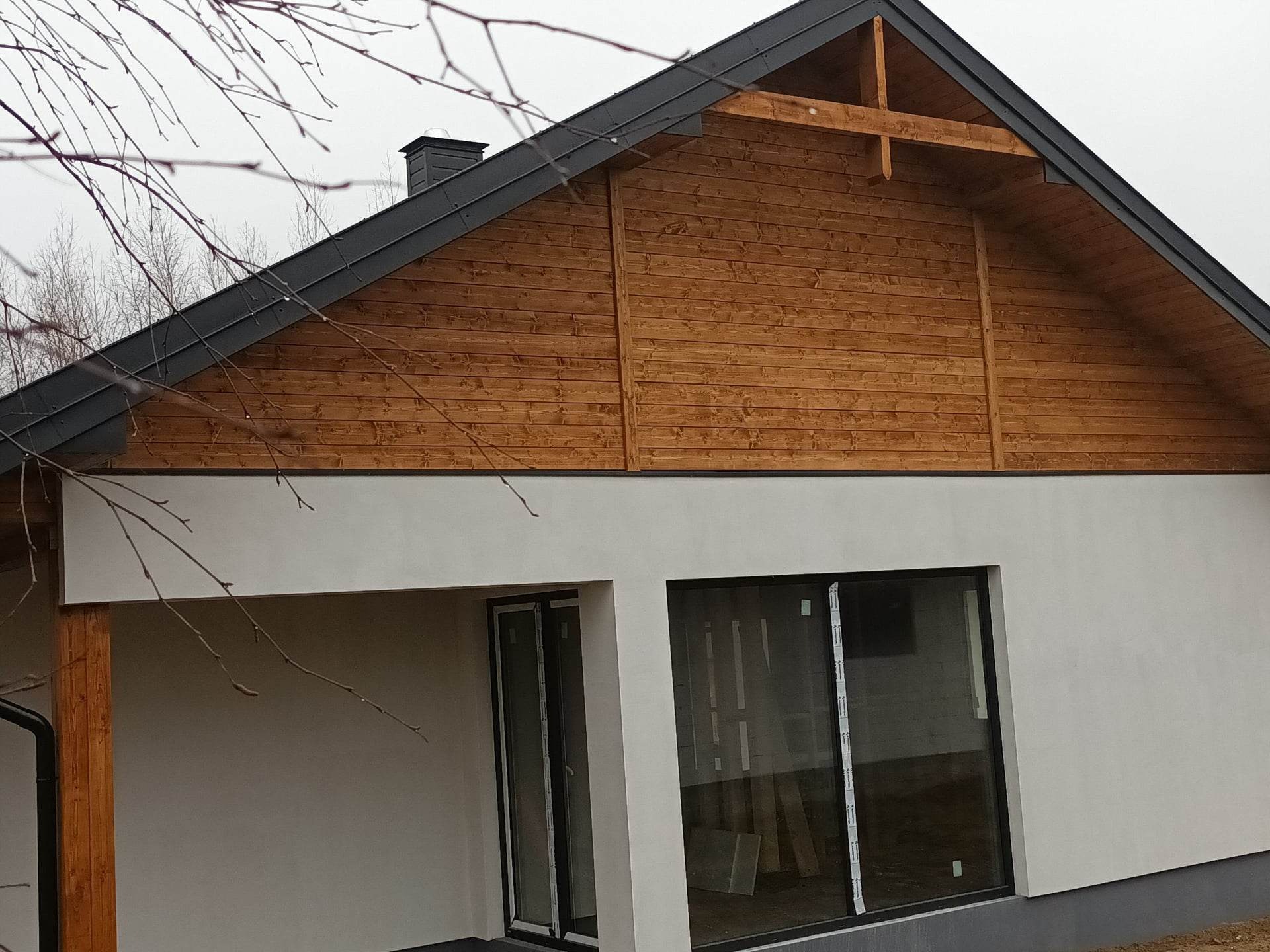 Fragment elewacji domu z drewnianą szczytową ścianą pod ciemnym dachem, z widocznymi dużymi oknami i minimalistyczną bryłą.