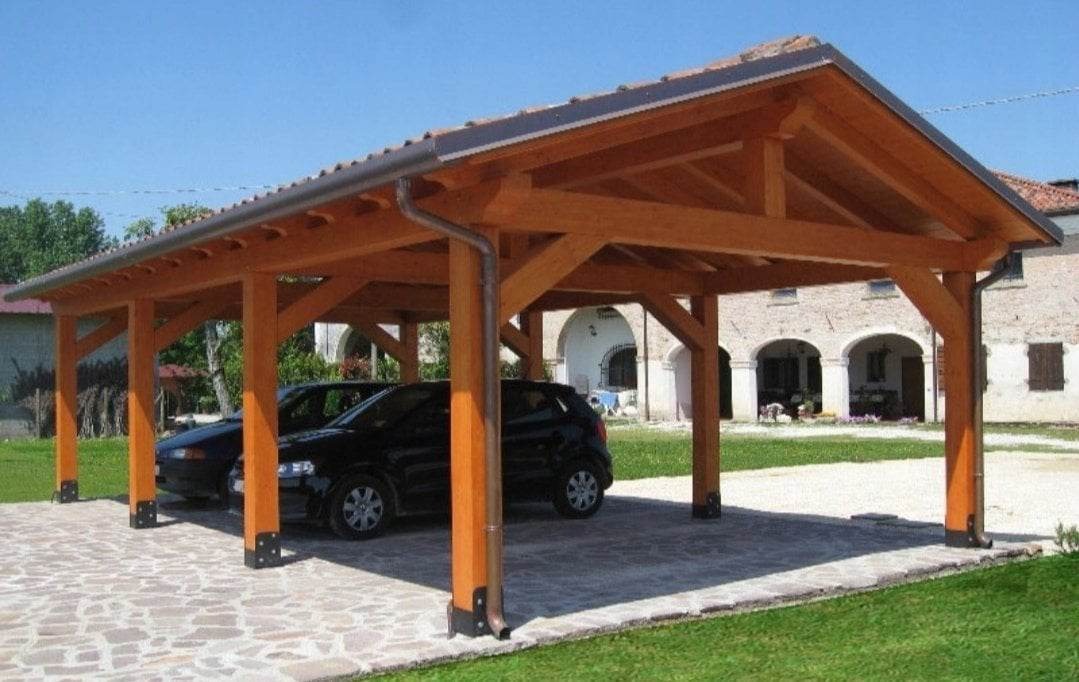 Drewniana wiata garażowa zadaszona dachówką, z dwoma samochodami zaparkowanymi na kamiennej nawierzchni, na tle budynku z arkadami.