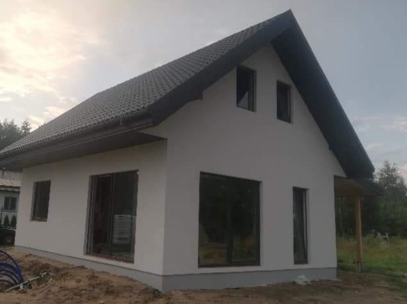 Surowy dom szkieletowy z ciemną dachówką i dużymi oknami, widok z boku na tle pochmurnego nieba i drzew.