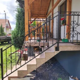 Zewnętrzne schody z terakoty z ozdobną, kutą, metalową balustradą w kolorze czarnym, prowadzące na zadaszony taras domu jednorodzinnego.