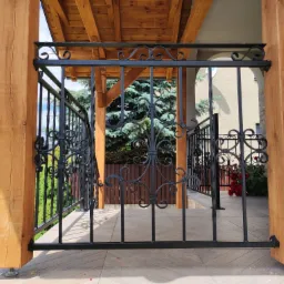 Czarna, metalowa balustrada balkonowa z ozdobnymi elementami, wsparta na drewnianych słupach, z widokiem na zielony ogród i elementy architektury domu w tle.