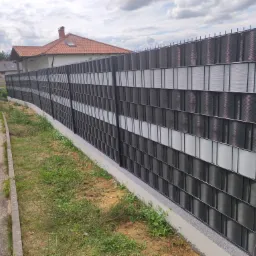 Długie ogrodzenie panelowe z poziomymi, naprzemiennymi pasami w kolorze grafitowym i białym, na betonowej podmurówce, z widocznym domem i ulicą w tle.