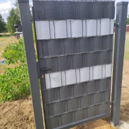 Nowoczesna brama wjazdowa z metalową ramą wypełnioną poziomymi panelami w odcieniach szarości i bieli, zamontowana na posesji z widoczną ziemią i zielenią w tle.