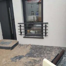 Czarna, pozioma krata okienna z metalowych prętów, zamontowana na elewacji budynku obok czarnych drzwi wejściowych, z widoczną kostką brukową i elementami budowlanymi na ziemi.