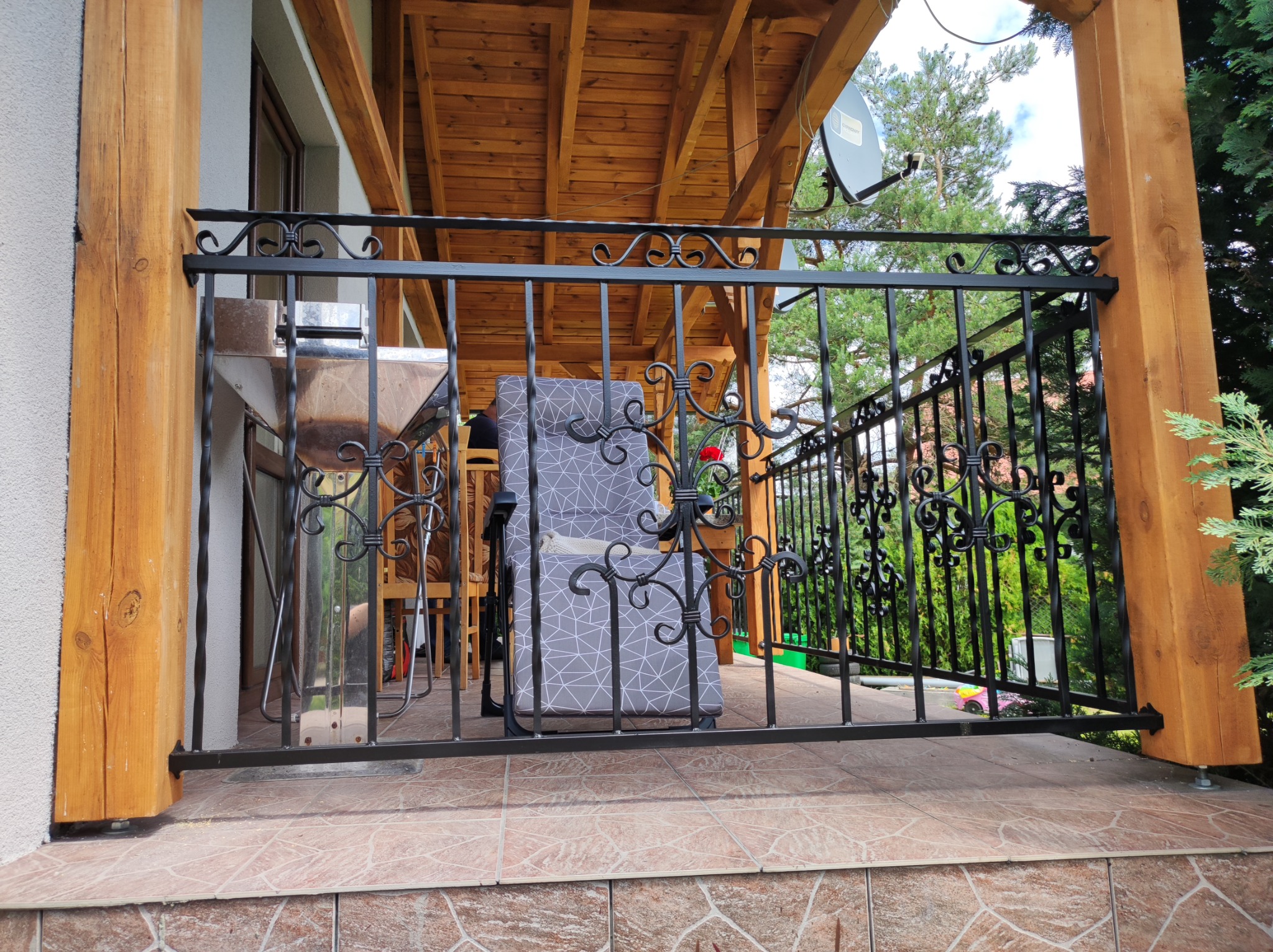 Czarna balustrada metalowa z dekoracyjnymi elementami na tarasie z drewnianą konstrukcją zadaszenia, widok na zieleń w tle.