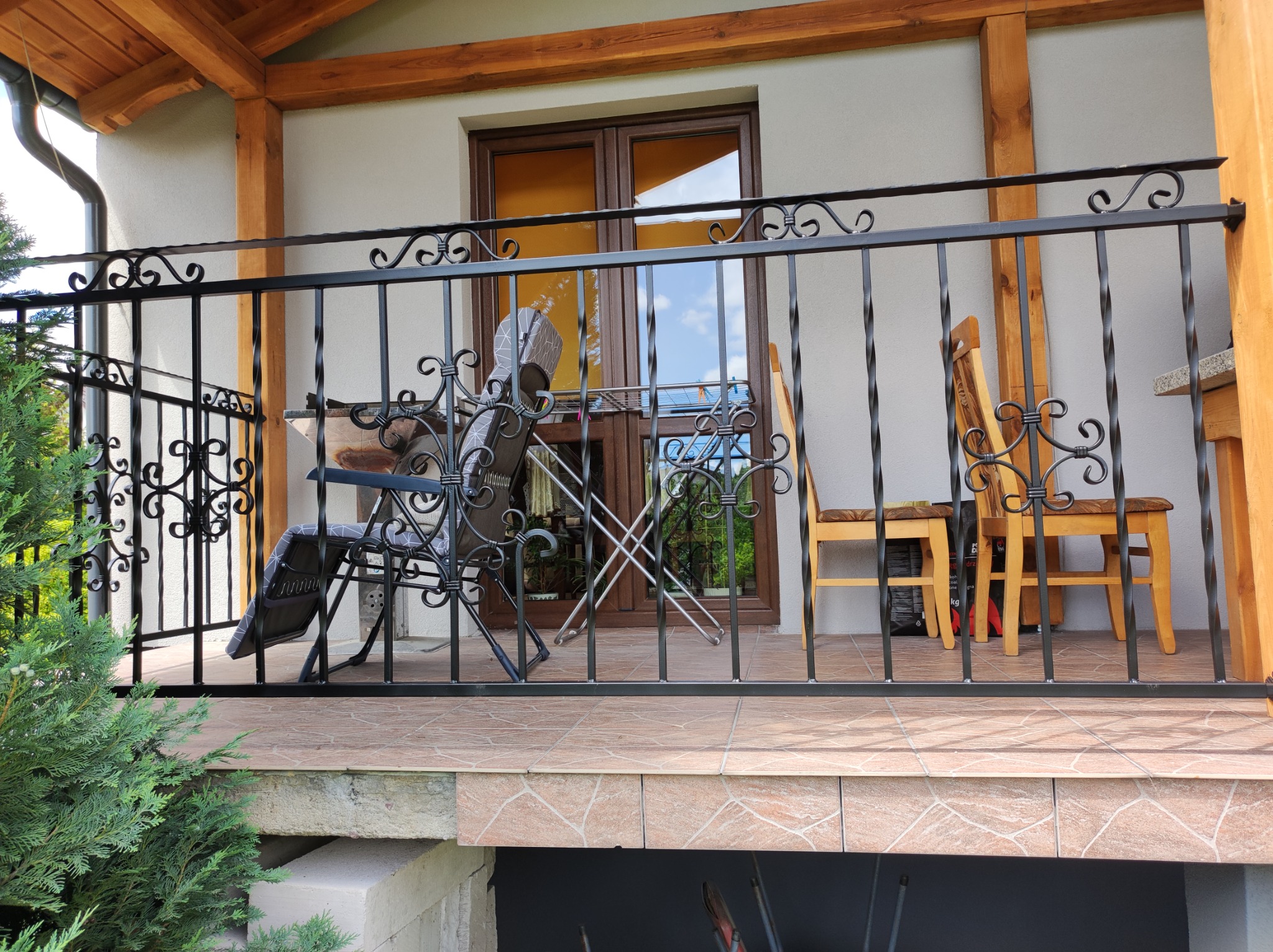 Czarna, metalowa balustrada balkonowa z ozdobnymi elementami, widok na taras z meblami ogrodowymi i roślinnością, jasna elewacja budynku z drewnianymi elementami konstrukcyjnymi.