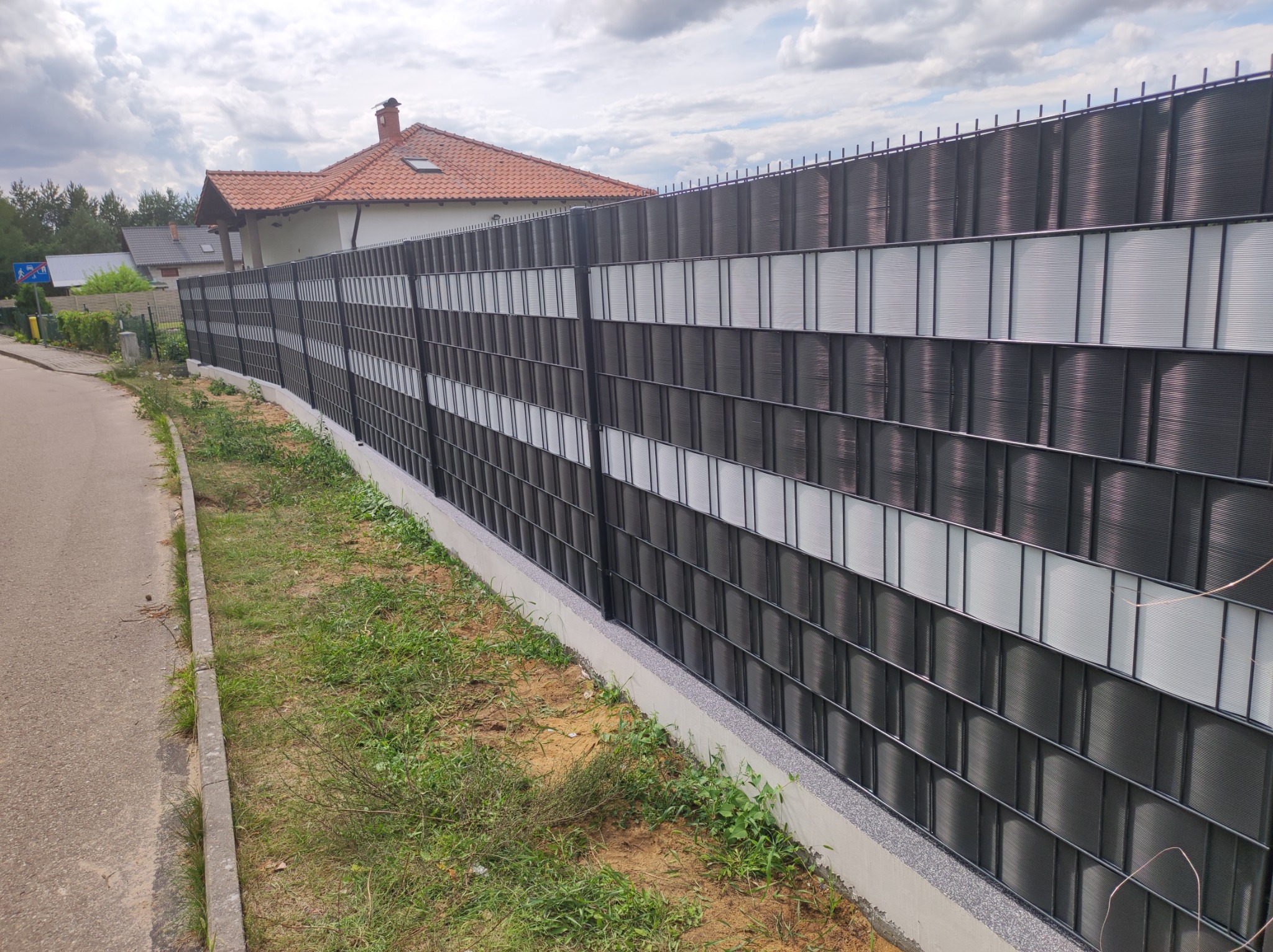 Długie ogrodzenie panelowe z poziomymi, naprzemiennymi pasami w kolorze grafitowym i białym, na betonowej podmurówce, z widocznym domem i ulicą w tle.