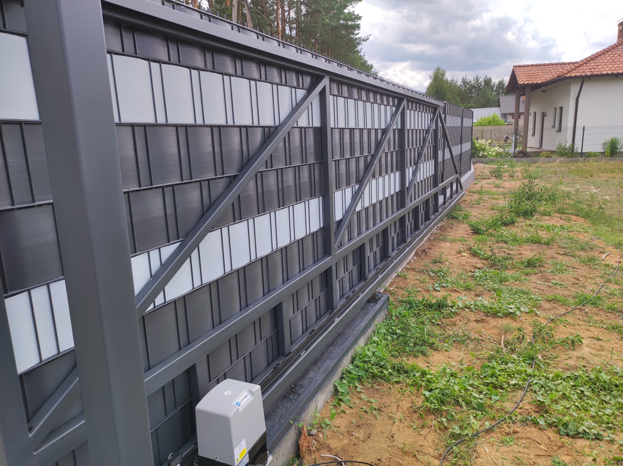 Przesuwna brama wjazdowa z metalową konstrukcją i wypełnieniem z paneli w kolorze szaro-białym, widoczny napęd elektryczny, trawa i dom w tle.
