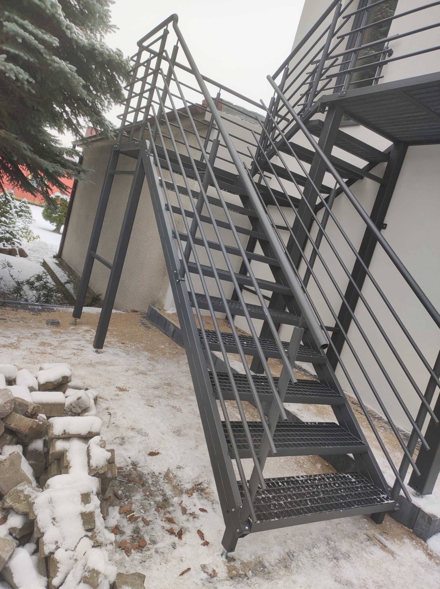 Zewnętrzne schody metalowe z kratowymi stopniami prowadzące na balkon, widoczne na tle ośnieżonego podwórka i budynku z jasną elewacją.