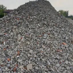 Stos pokruszonego betonu i cegieł na placu budowy, widoczny fragment dachu domu w tle.