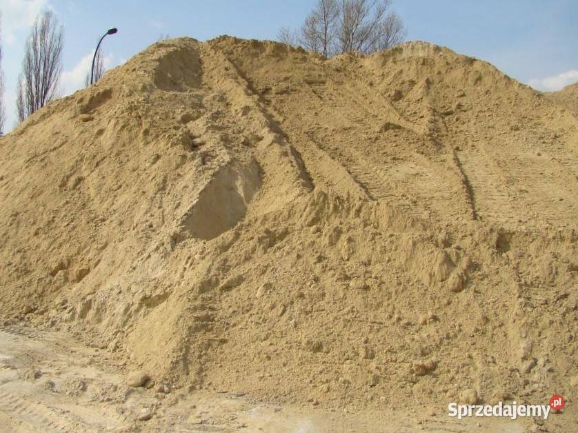 Duża pryzma piasku z widocznymi śladami opon pojazdu budowlanego, ustawiona na placu budowy, w tle drzewa bez liści i słup oświetleniowy.