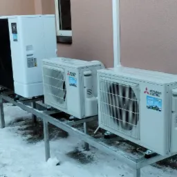 Zewnętrzne jednostki klimatyzacji i pompa ciepła Mitsubishi Electric zamontowane na metalowej konstrukcji wsporczej, widoczne na tle elewacji budynku w zimowej scenerii.