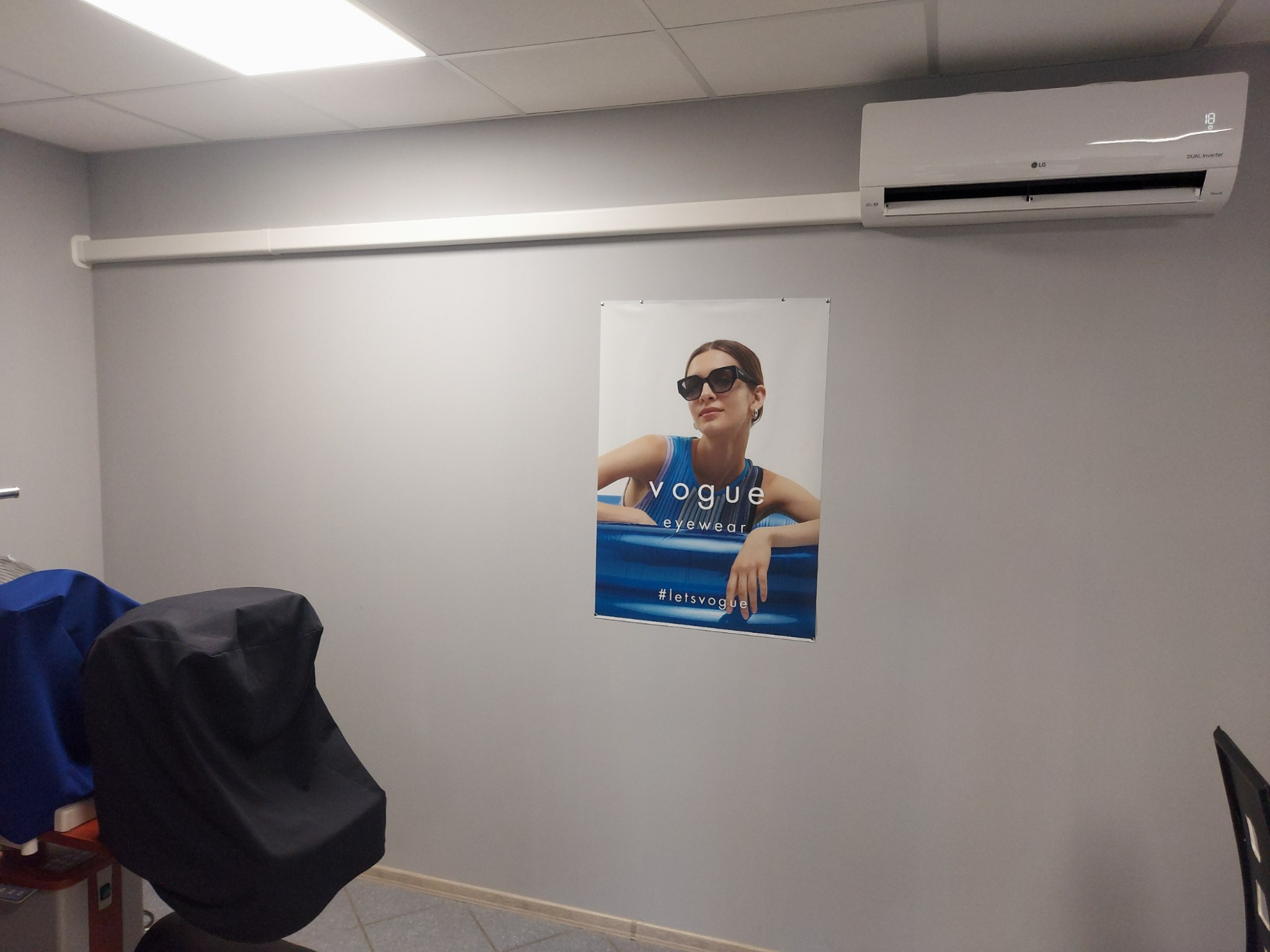 Biały klimatyzator LG Dual Inverter zamontowany na szarej ścianie nad plakatem Vogue Eyewear w pomieszczeniu z kasetonowym sufitem.