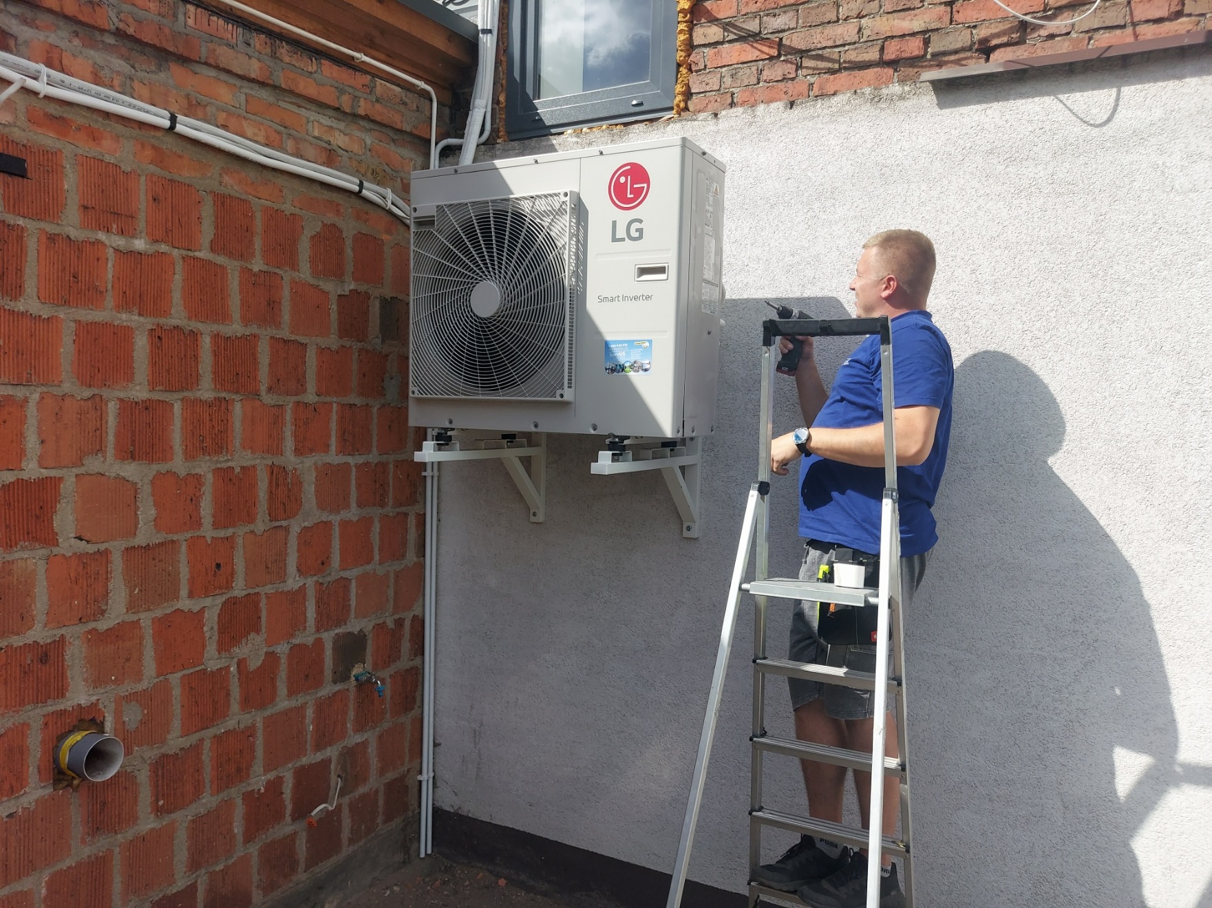 Monoblok zewnętrzny klimatyzatora LG Smart Inverter zamontowany na białej ścianie, instalator na drabinie z wiertarką, widoczne rury instalacyjne na ceglanej ścianie budynku.
