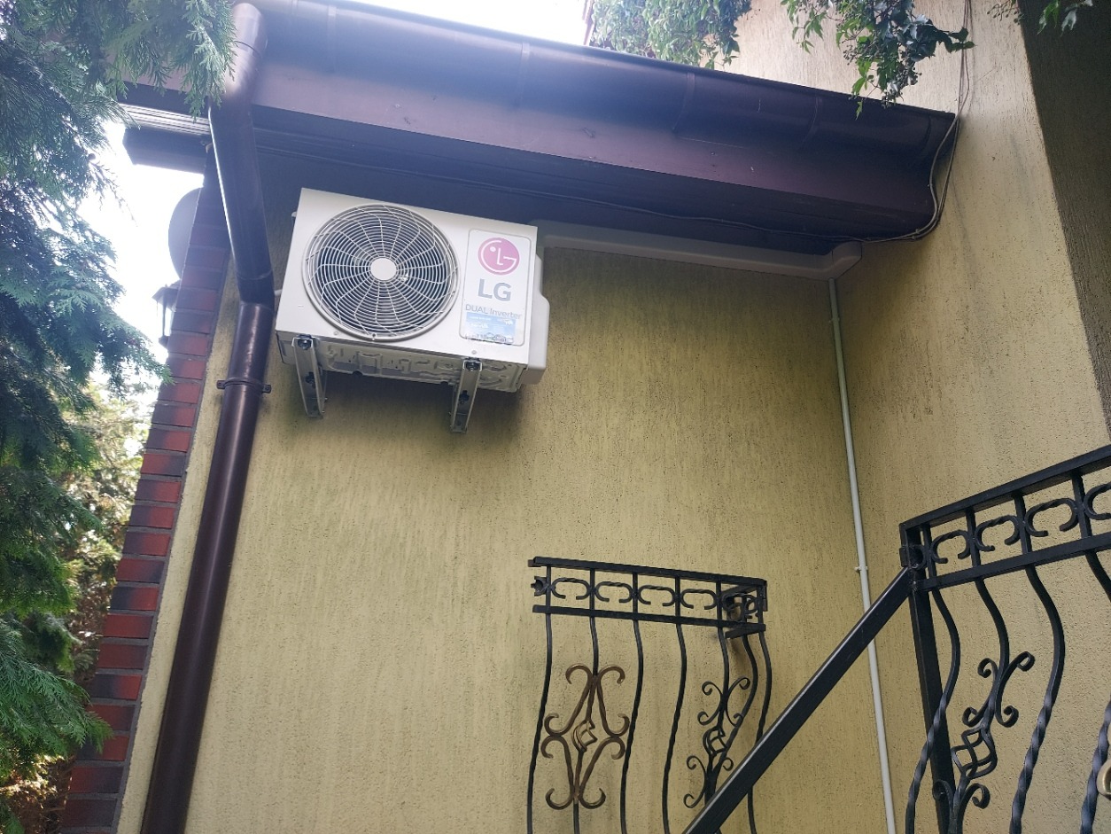 Zewnętrzna jednostka klimatyzacji LG zamontowana na elewacji budynku z widocznymi elementami orynnowania i fragmentem dekoracyjnej, metalowej balustrady schodów.