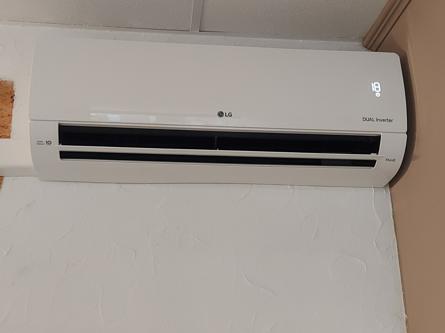 Zamontowany biały klimatyzator LG Dual Inverter z wyświetlaną temperaturą 18 stopni Celsjusza na ścianie o fakturze baranka.