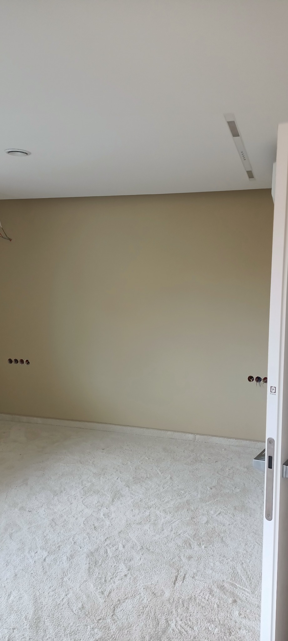 Pokój w stanie deweloperskim z beżową ścianą, widoczne gniazdka elektryczne, wylewka na podłodze i białe drzwi przesuwne.