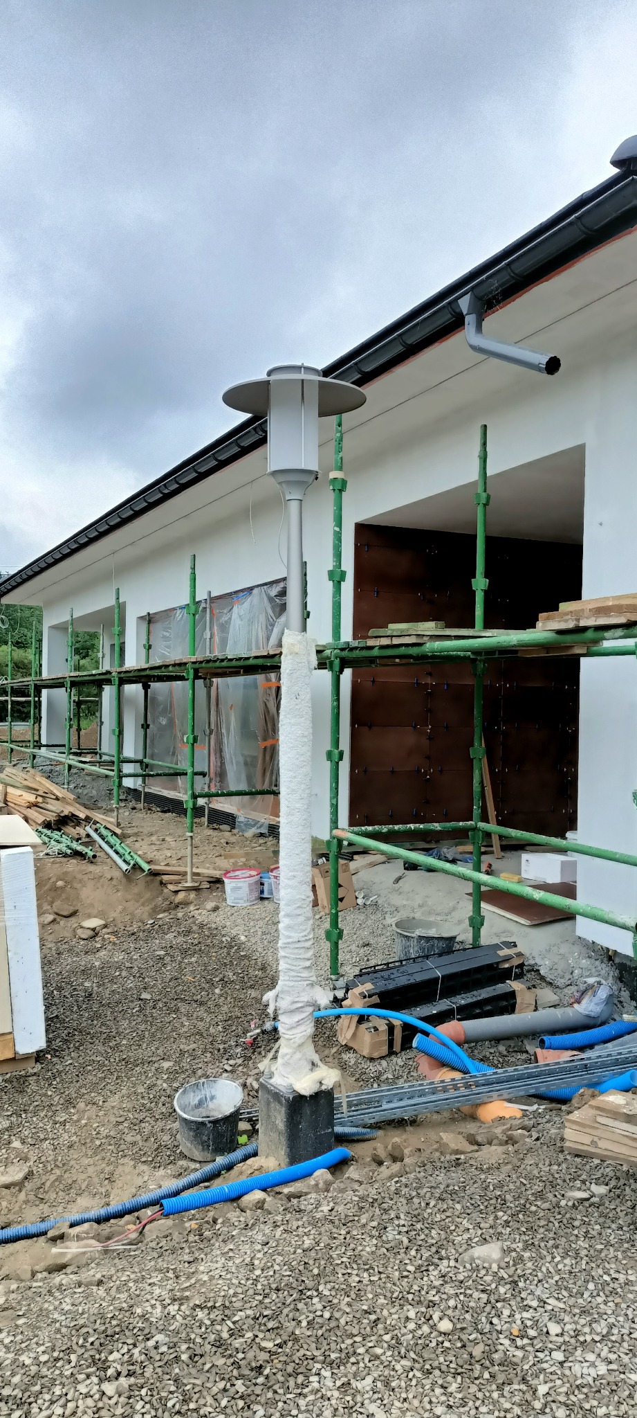 Budynek w trakcie prac elewacyjnych z widocznymi rusztowaniami, zabezpieczonym słupem oświetleniowym i przewodami w ziemi.