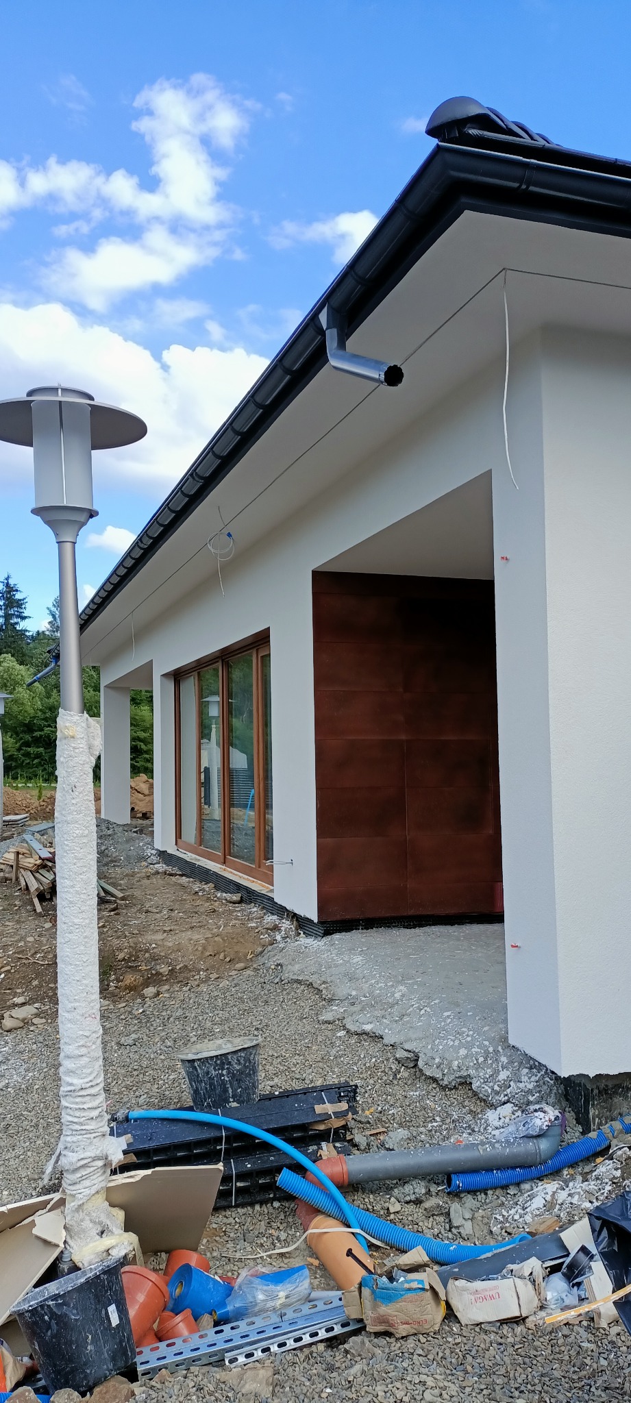 Nowoczesny dom jednorodzinny w Nowym Sączu z białymi ścianami zewnętrznymi i brązową, dużą bramą garażową, widoczne elementy elewacji oraz lampa zewnętrzna, teren wokół w trakcie prac wykończeniowych.