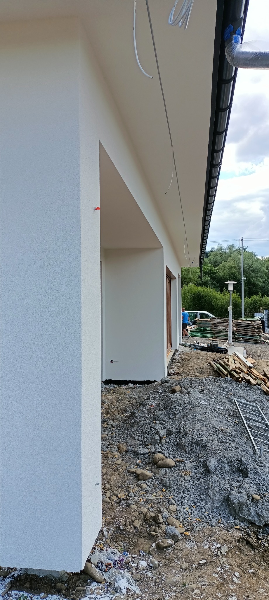 Świeżo otynkowana elewacja budynku jednorodzinnego w kolorze białym, widoczny fragment dachu z rynną oraz teren wokół budynku w trakcie prac wykończeniowych z gruzem i materiałami budowlanymi.
