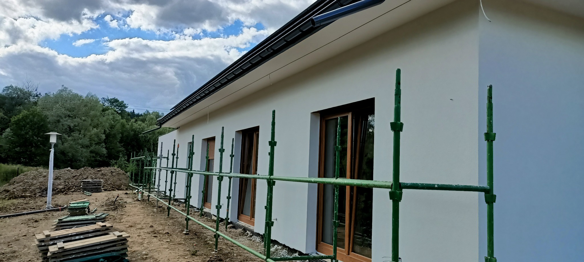 Nowo otynkowana, biała elewacja domu jednorodzinnego z drewnianymi oknami, widoczna z poziomu rusztowania w trakcie prac wykończeniowych.