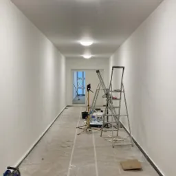 Długi korytarz w trakcie przygotowania do malowania: podłoga zabezpieczona folią, ściany świeżo pomalowane na biało, widoczne drabiny, narzędzia malarskie i rolka kabla.