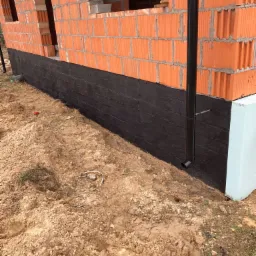Fragment ściany budynku w trakcie budowy: czerwony mur z pustaków ceramicznych, zabezpieczona czarną izolacją fundamentowa ściana oraz fragment ocieplenia ze styropianu.