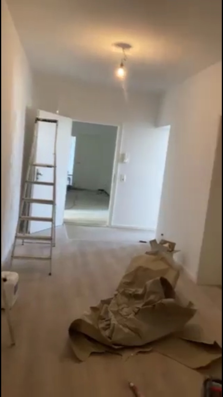 Wnętrze pokoju w trakcie malowania, z białymi ścianami, rozłożonym papierem ochronnym na podłodze, drabiną opartą o ścianę i wiszącą żarówką