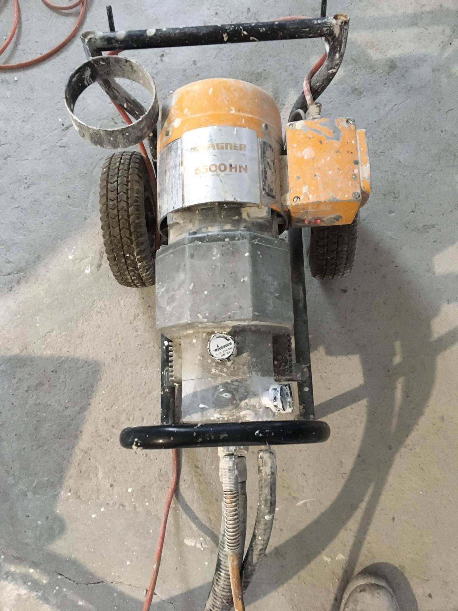 Używany agregat malarski Wagner 6500 HN z widocznymi śladami farby na obudowie, kołach i przewodach, stojący na betonowej podłodze z odpryskami farby.