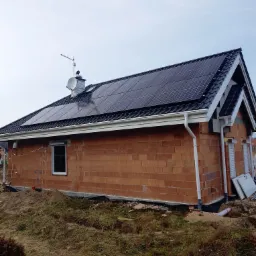 Dom w Łodzi z panelami fotowoltaicznymi na dachu, widoczna antena satelitarna i komin. Budynek w trakcie budowy, z surowymi ścianami z cegły.
